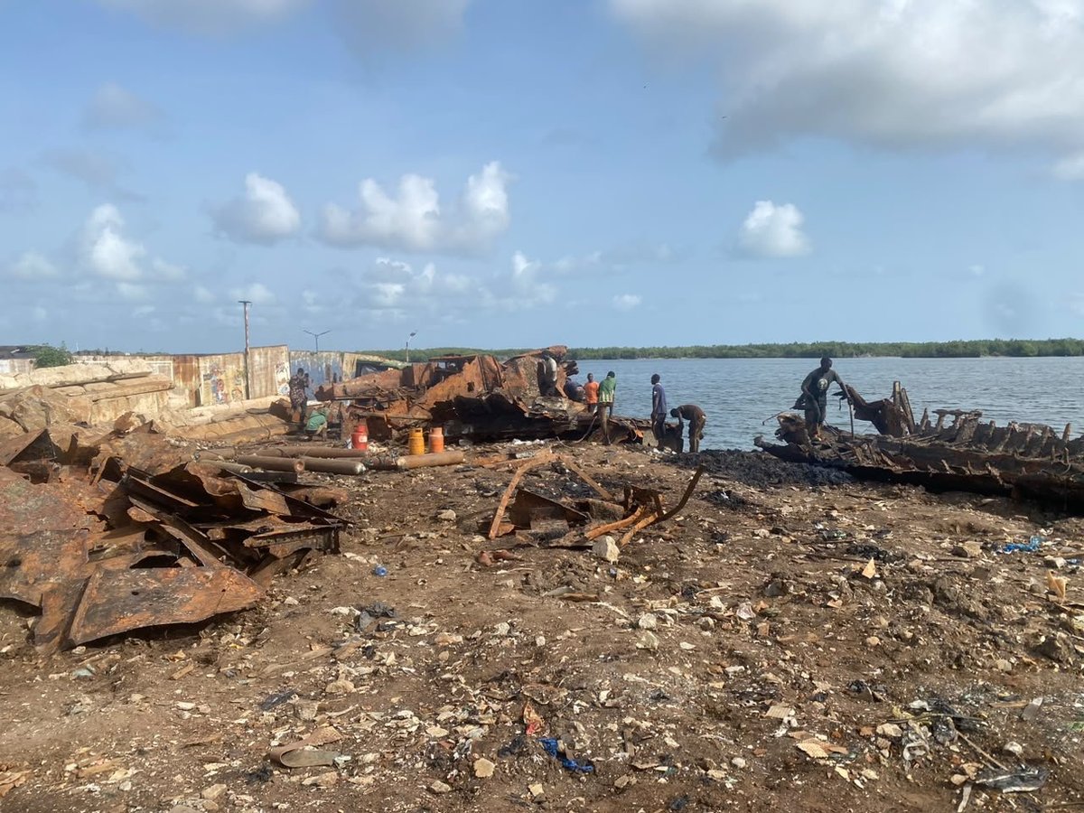 Industrial waterfront with heavy machinery, scattered metal debris, and workers on a rocky beach beside calm water.
