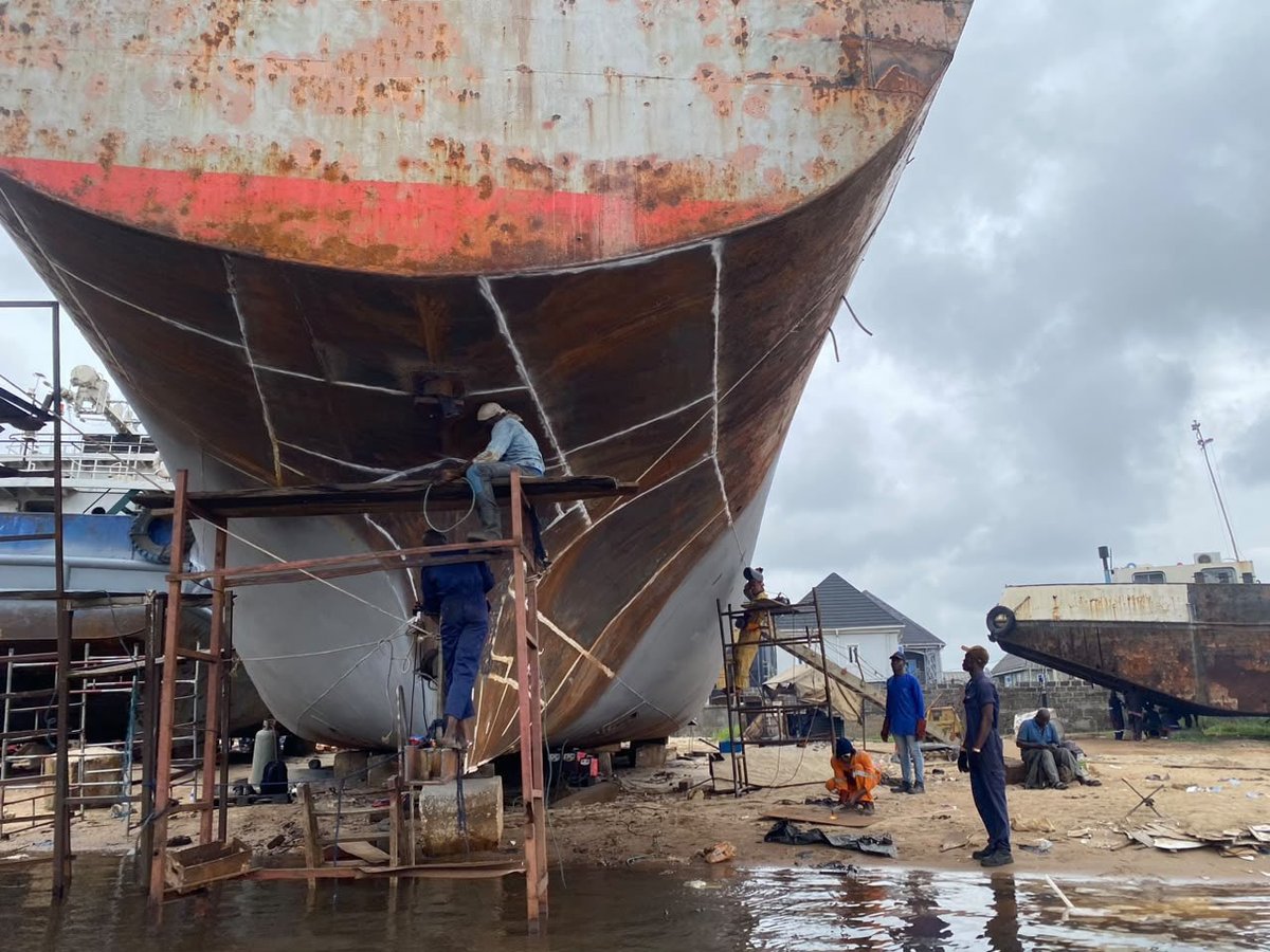 Workers in blue uniforms dismantling a large rusted ship hull in a shipyard under cloudy skies.