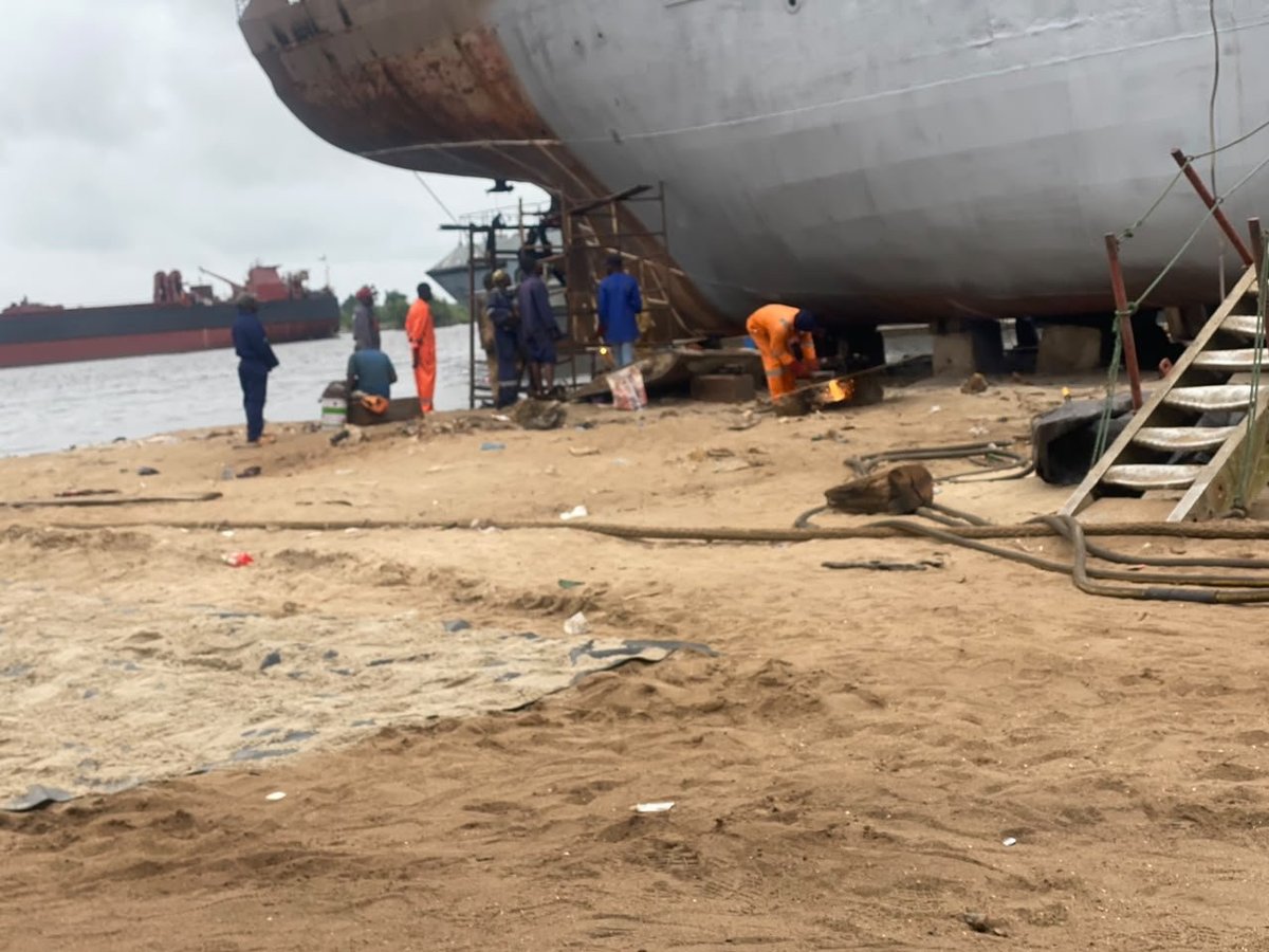 Large ship under repair on sandy beach with workers in safety gear standing nearby during maintenance work.
